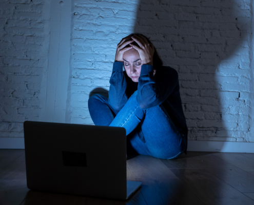 Image of a teen girl sitting in a dark room on the floor in front of the laptop holding her head in worry