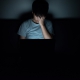 Image of a young boy sitting in the dark in front of a laptop and holding his head in worry