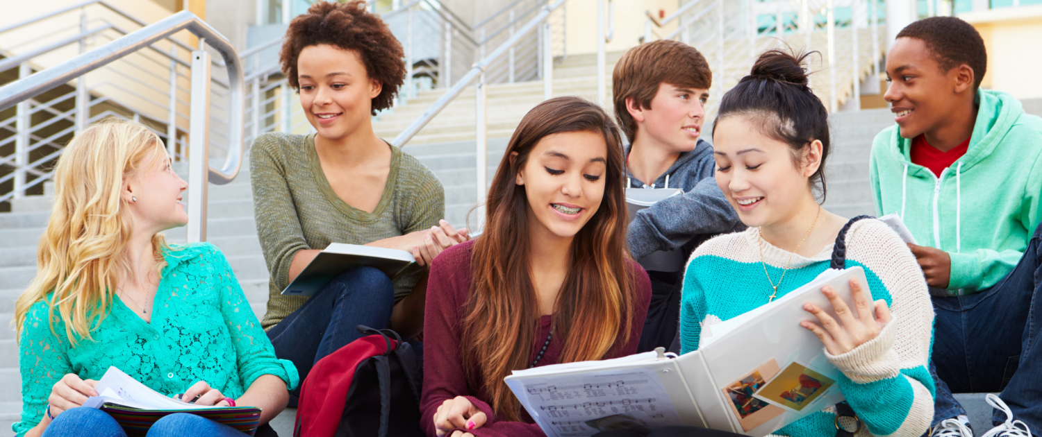 Image of a group of teens (4 girls and 2 boys) sitting on stairs in front of the school. 2 girls are reading some notes from the notebook and smiling, 2 boys are facing each other and smiling and the remaining 2 girls are facing each other and smiling.