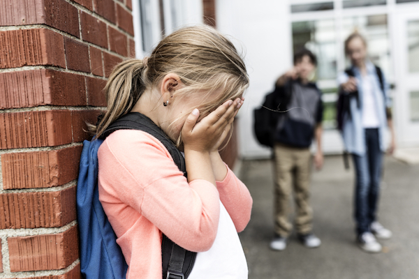 Image of a young girl against the wall and crying in her palms while two boys in the blurry background are pointing at her