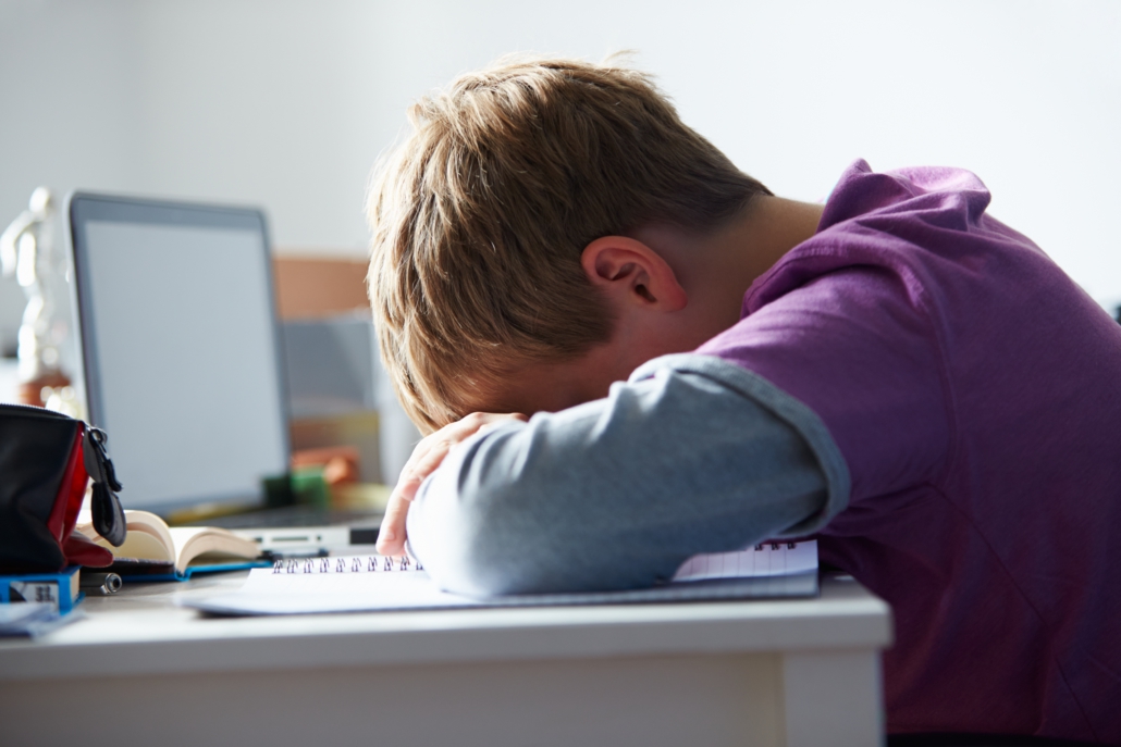 Image of a young boy crying over his arms while sitting on his desk in front of a computer