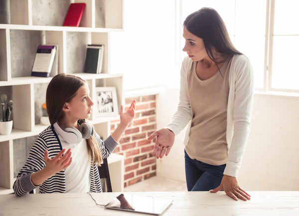 Image of a teen girl sitting down and possibly arguing with her mother who is standing next to her pointing to the iPad on the desk