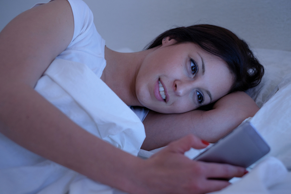 Image of a smiling young woman lying in bed and looking at her smartphone